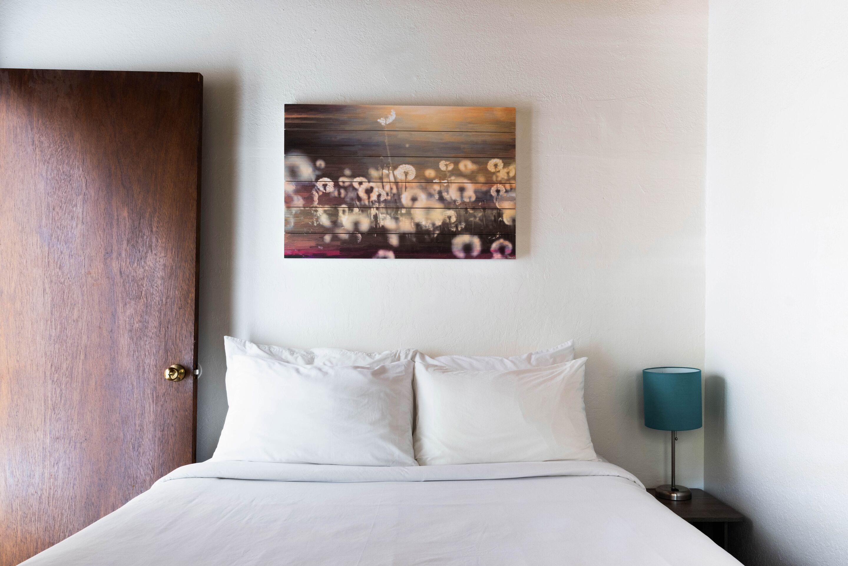 Een hotelkamer met een houten deur, een bed met witte lakens, een nachtkastje met een lamp en een kunstwerk aan de muur.