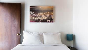 A hotel room with a wooden door, a bed with white linens, a nightstand with a lamp, and a wall-mounted artwork.