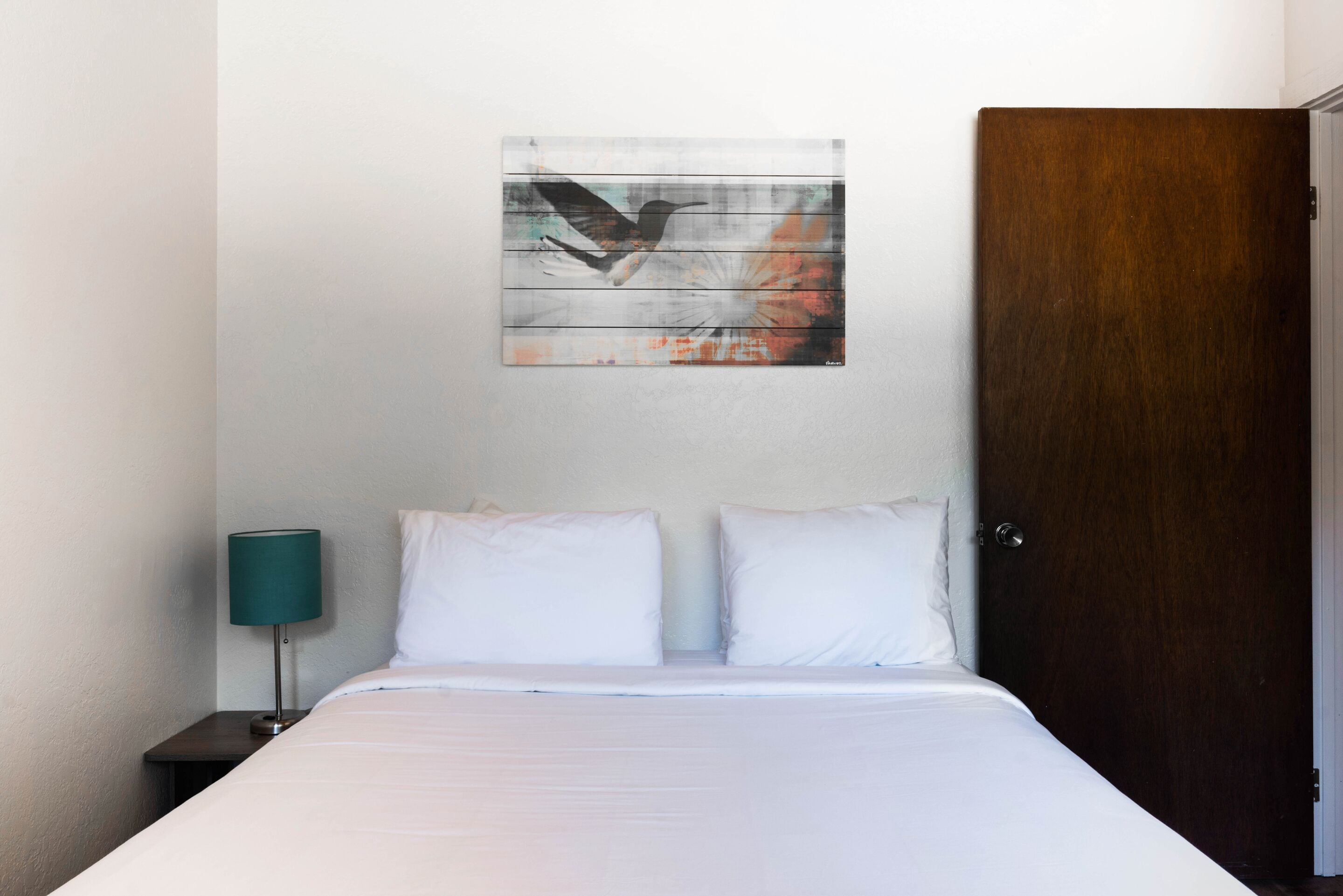 A hotel room with a bed, two pillows, a teal lamp, and a wooden door.