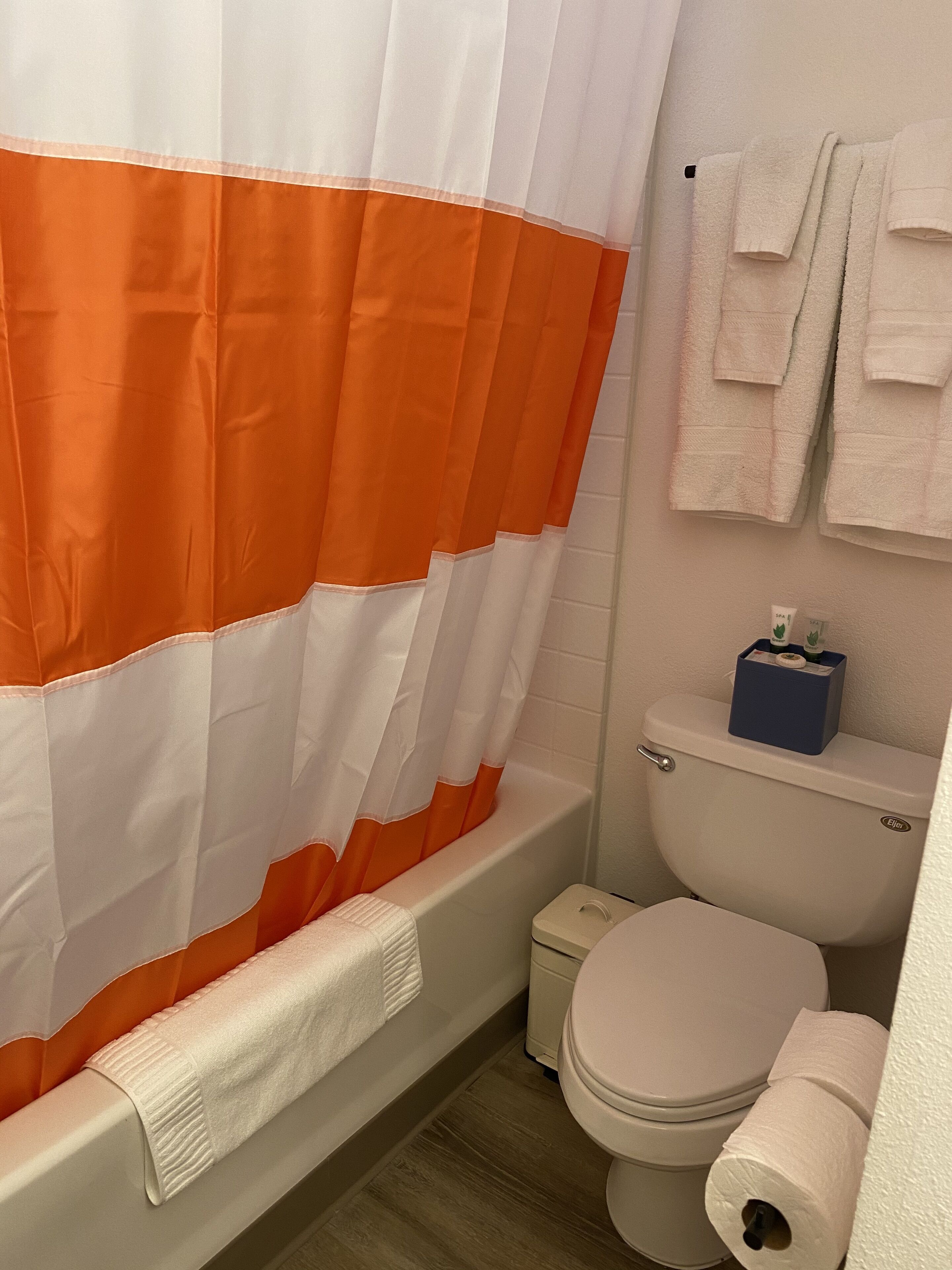 A bathroom with a shower curtain, a toilet, a small trash bin, and a towel rack with towels.