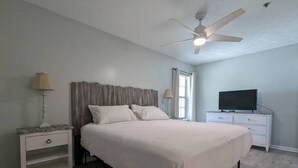 Ein Schlafzimmer mit einem Bett, einem Nachttisch, einer Lampe, einem Deckenventilator, einem Fernseher und einem Fenster.