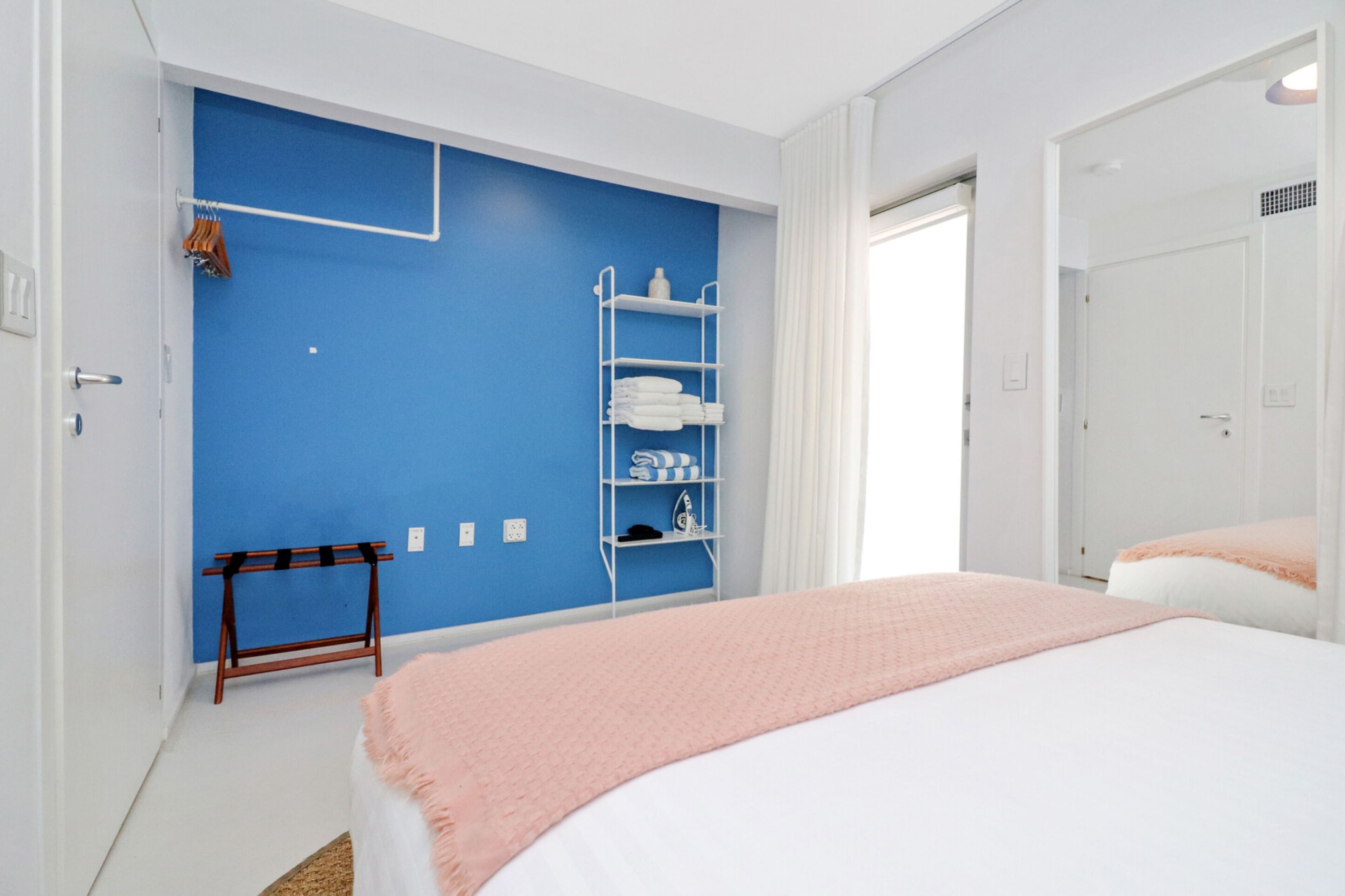 Un dormitorio con pared azul de acento, un armario blanco, una cama con manta rosa y un estante con toallas y artículos de aseo.