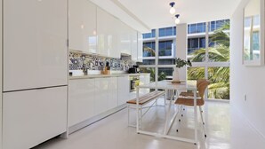 Une cuisine moderne avec des meubles blancs, une table à manger avec des chaises et une vue sur les palmiers à travers de grandes fenêtres.