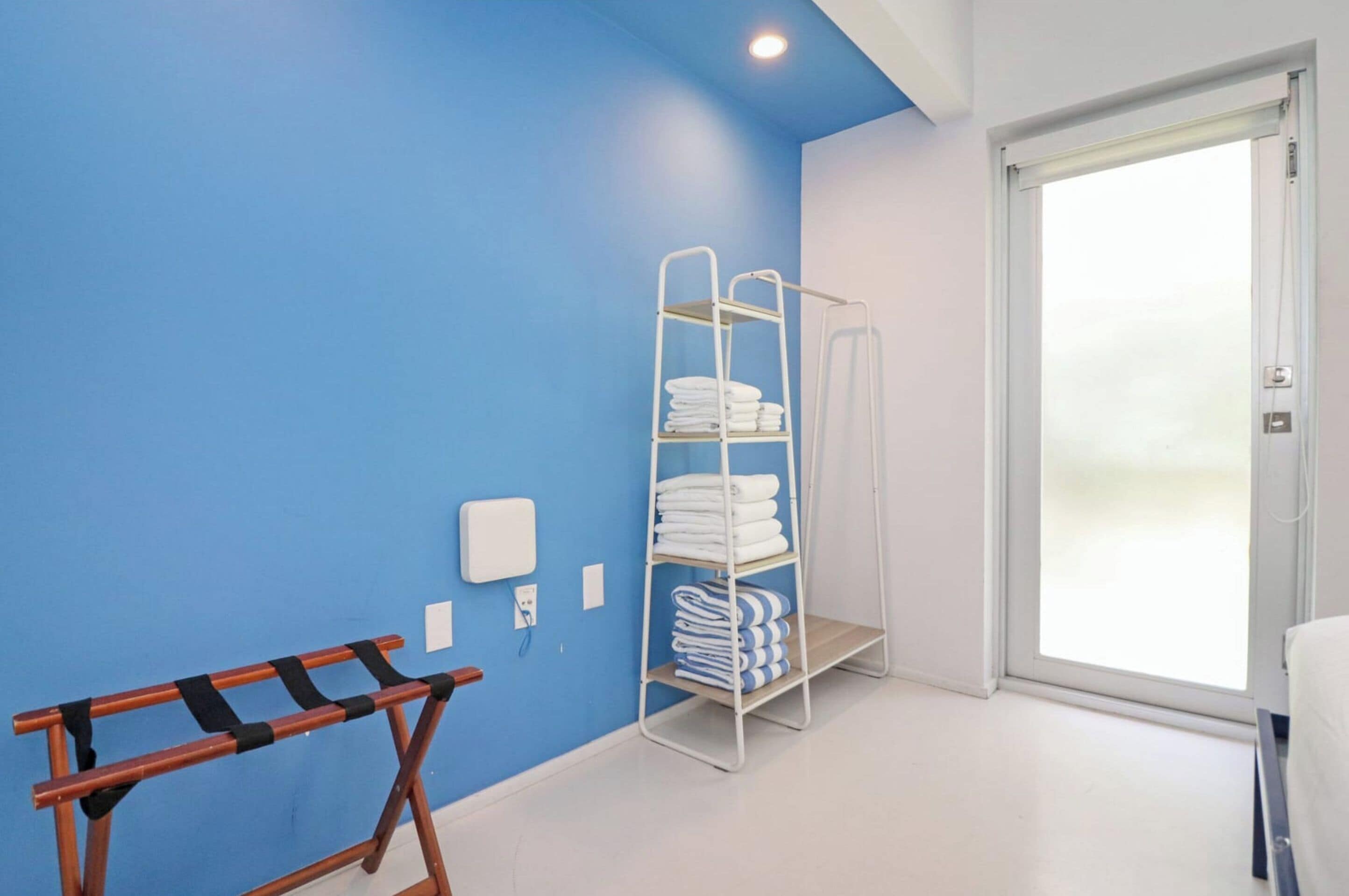 Hotelkamer met een blauwe wand, een metalen plank met opgevouwen handdoeken, een houten bankje en een deur naar een lichte ruimte.