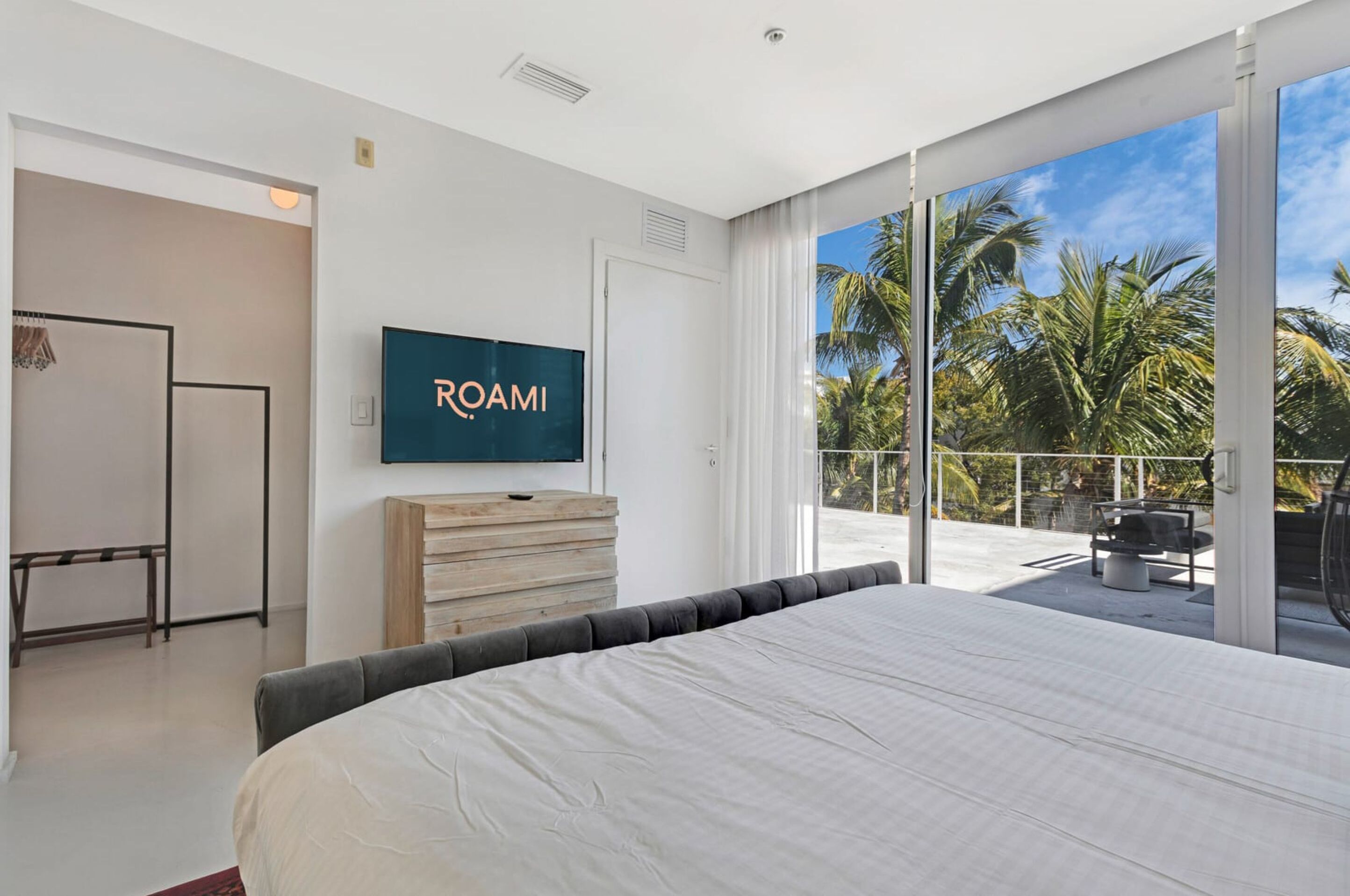 Un dormitorio moderno con una cama grande, un televisor de pantalla plana, un aparador de madera y un balcón con vista a palmeras.