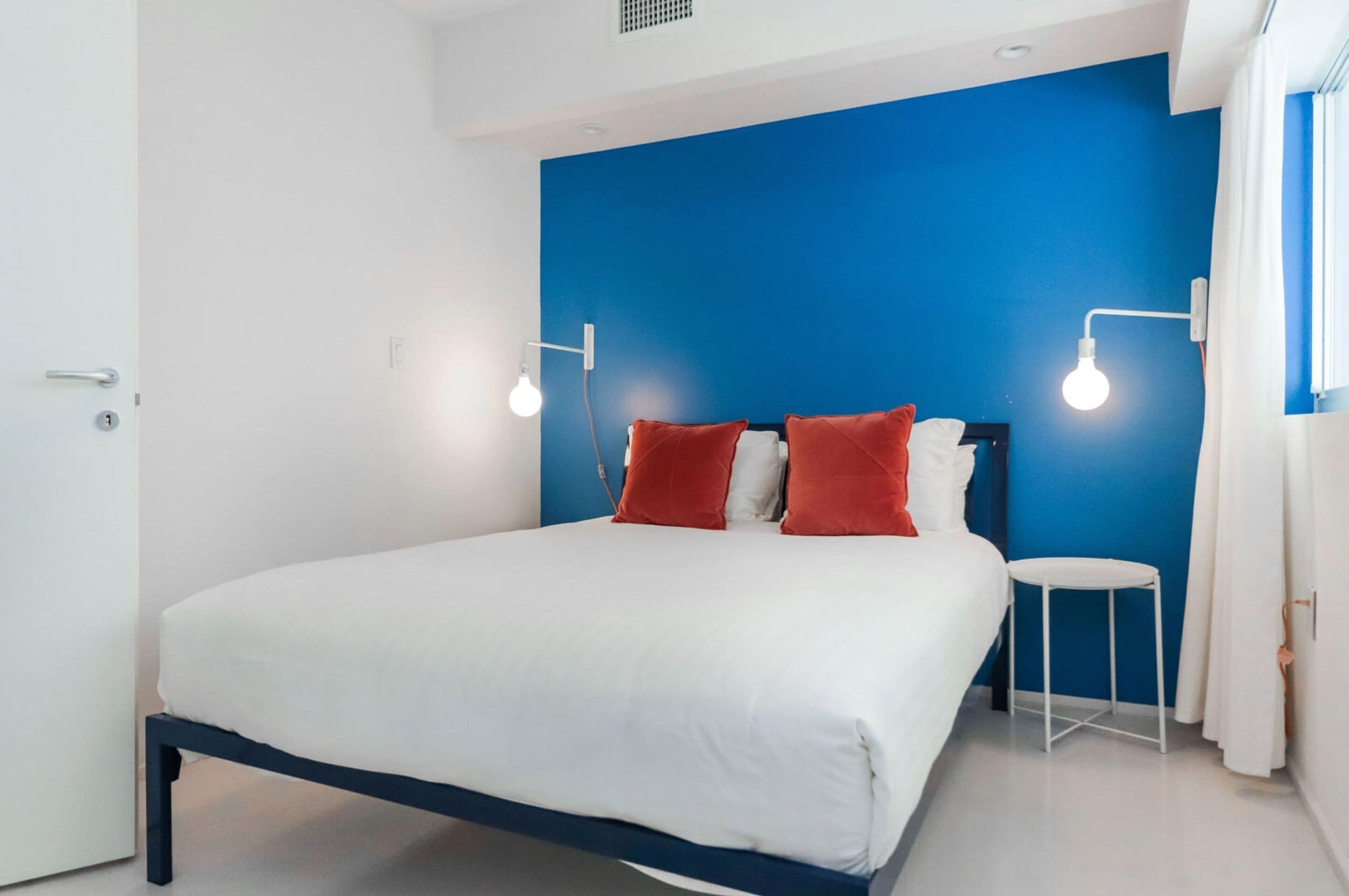 A bedroom with a bed, two bedside lamps, a small white stool, and a window with curtains.