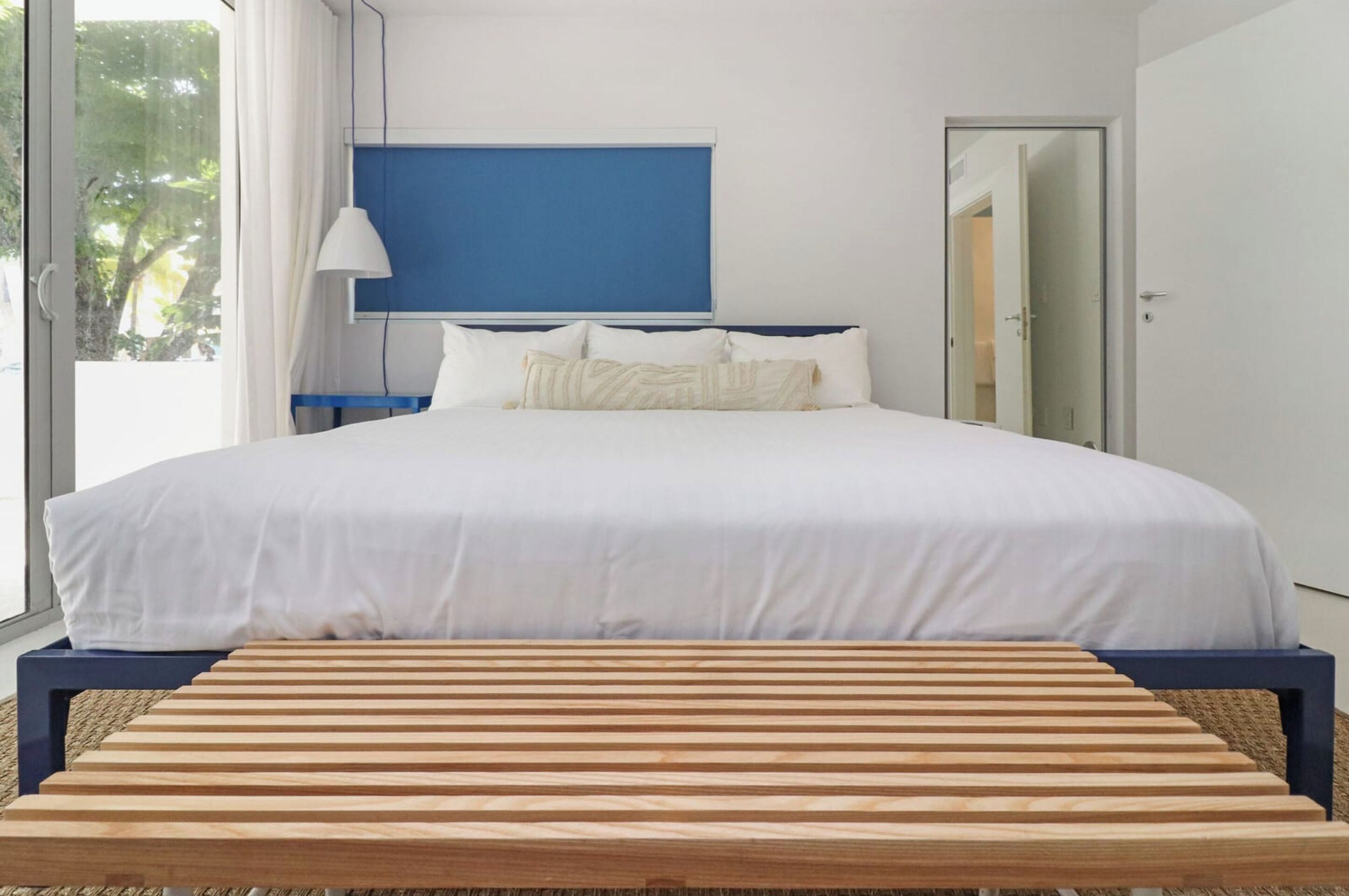A modern bedroom with a large bed, a wooden bench, and a view of the outdoors.