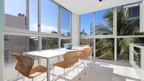 Une pièce lumineuse, baignée de soleil, avec de grandes fenêtres, une table blanche, des chaises en osier et un bol posé sur la table.