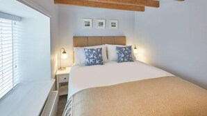 A bedroom with a bed, bedside lamps, a window with blinds, and a wooden ceiling beam. - Amber Cottage (Whitby)