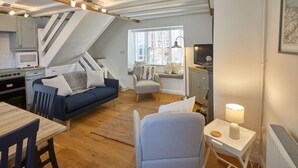 A cozy living room with a sofa, a dining table, and a chair. There's a kitchen area with a microwave and a stove. A window provides natural light, and a lamp is on a side table. - Amber Cottage (Whitby)