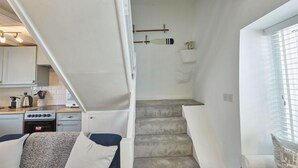 A compact living space with a kitchenette, a staircase, and a window with blinds. - Amber Cottage (Whitby)