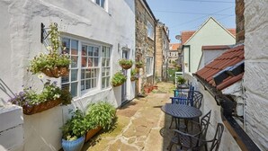 A narrow courtyard with stone flooring, a small table and chairs, and hanging flower baskets on the walls. - Amber Cottage (Whitby)