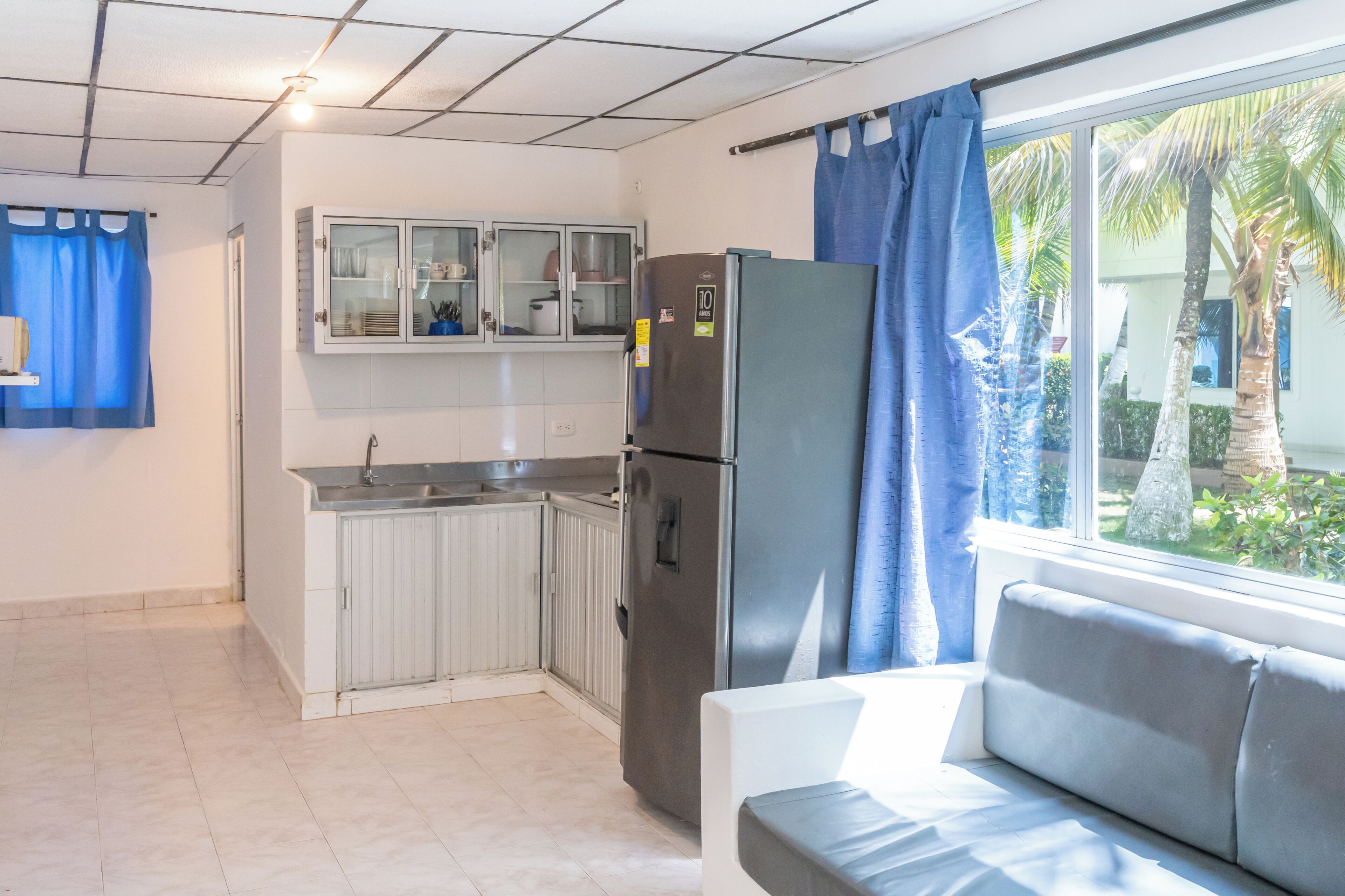 A compact kitchen with a refrigerator, a window with curtains, and a view of palm trees.