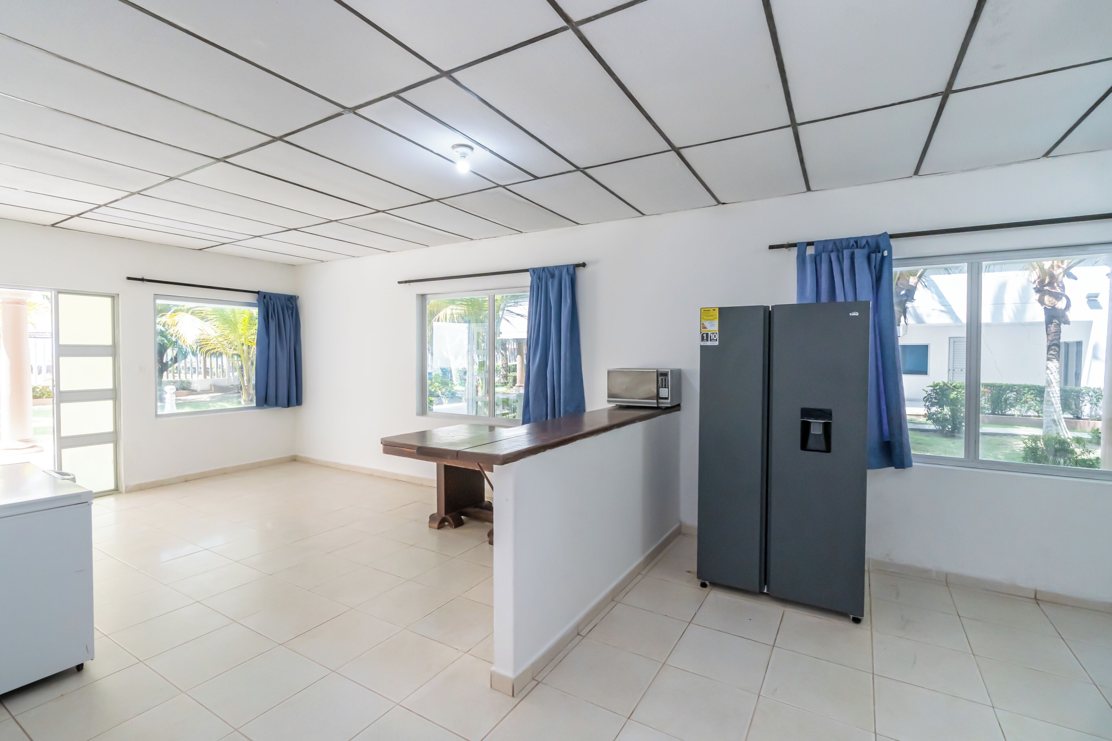 A spacious kitchen with a refrigerator, microwave, and a window with blue curtains.