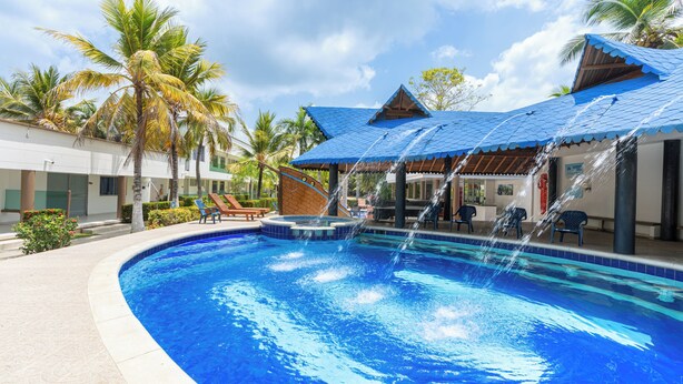 A swimming pool with water features, palm trees, and a covered area with chairs. - Cabañas La Fragata Coveñas (Coveñas)
