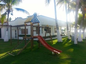A playground with a slide and climbing structures, surrounded by palm trees and a white building. - Cabañas La Fragata Coveñas (Coveñas)