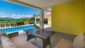 Un balcon agrémenté de meubles en osier, une table avec des fruits et une vue sur une piscine et la verdure.