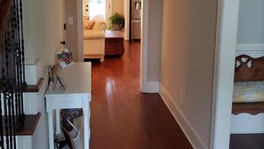 A hallway with a wooden floor, a white wall, and a door leading to a living room with a sofa and a table. - Augusta Golf Course Rental Home MASTERS (Evans)