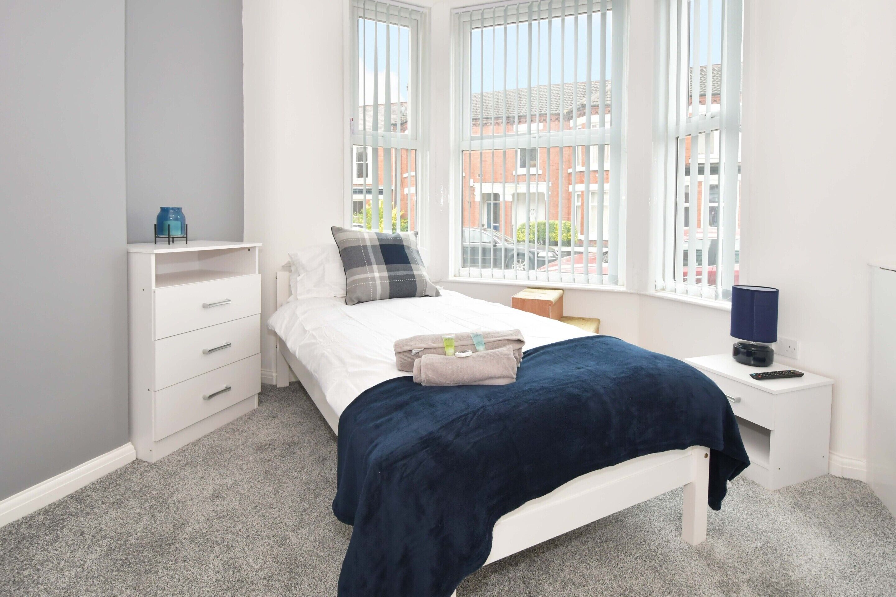 Quarto com cama, mesa de cabeceira, cômoda branca e janela com vista para edifícios.