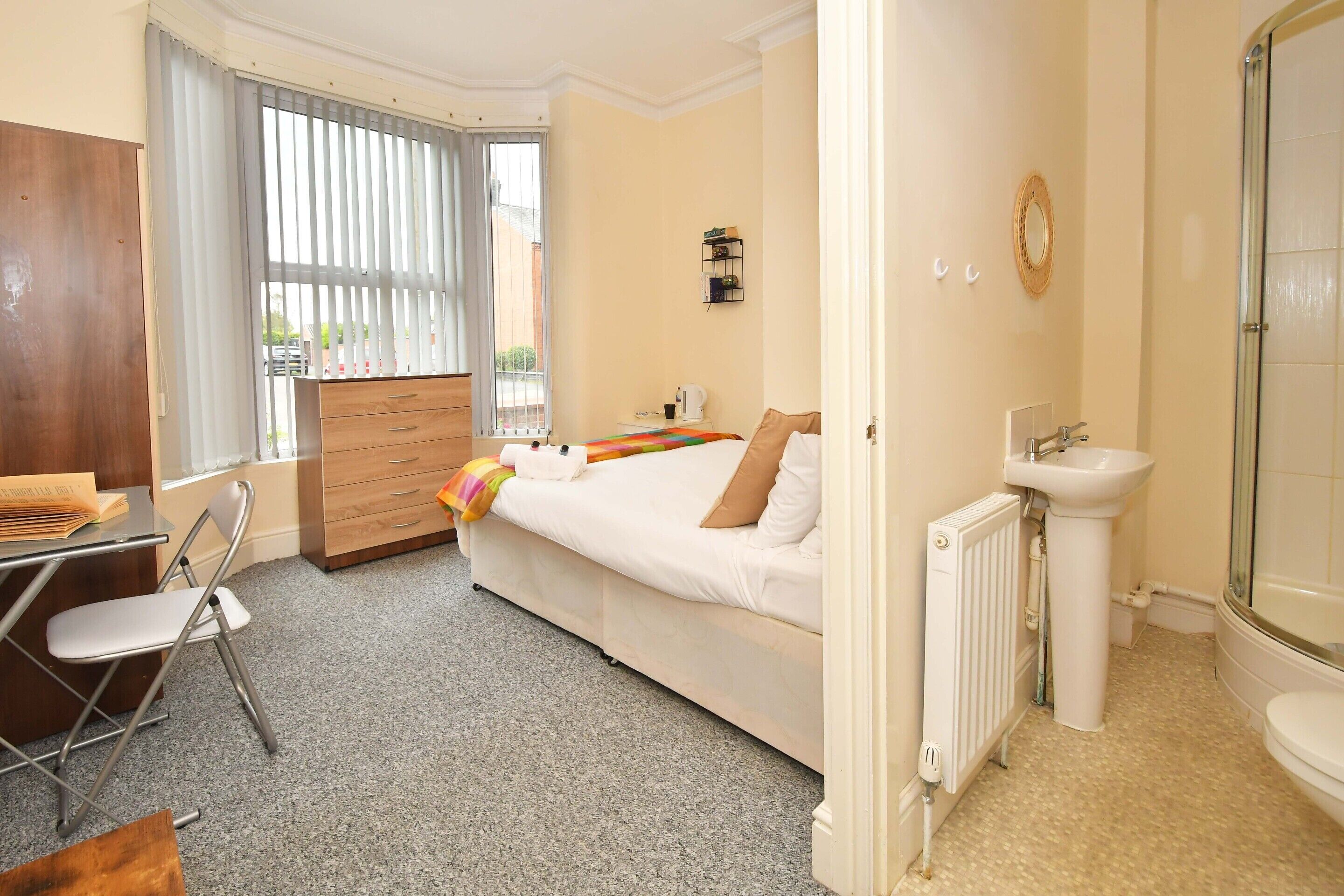 A bedroom with a bed, a desk, a chair, a dresser, a radiator, a sink, and a window with curtains.