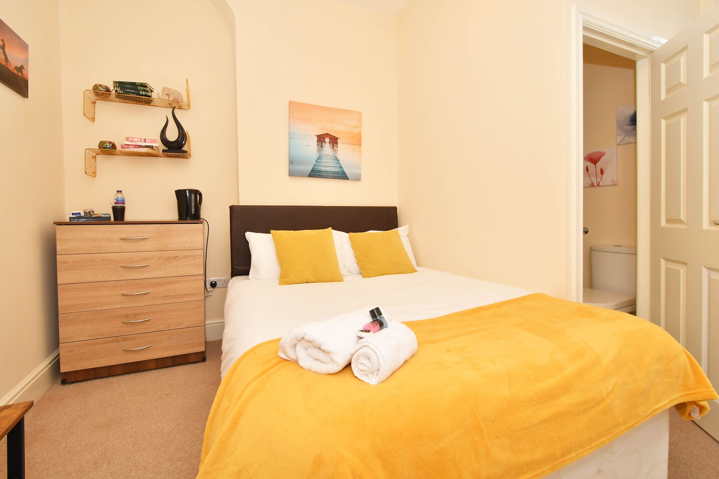 A neatly made bed with a yellow blanket, a wooden dresser with drawers, a shelf with books and decorative items, and a painting of a pier on the wall.