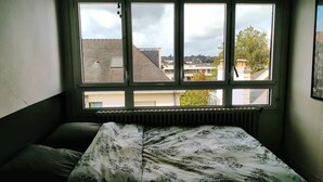 A bedroom with a bed, a radiator, and a window with a view of buildings and trees. - Connected studio with open view - CENTRE-VILLE (Nantes)