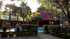 Un edificio rojo con tejado de tejas, rodeado de abundante vegetación y una piscina.