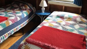 A room with two beds, each with a colorful quilt, a small table with a lamp, and a window with curtains. - Airy Wooden Beach House with Big Private Deck, close to Atlantic Ocean Beach or gentler bay swimming.  (Fire Island)