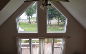 A room with a sloped ceiling, two large windows, a ceiling fan, and a view of a grassy area with trees.