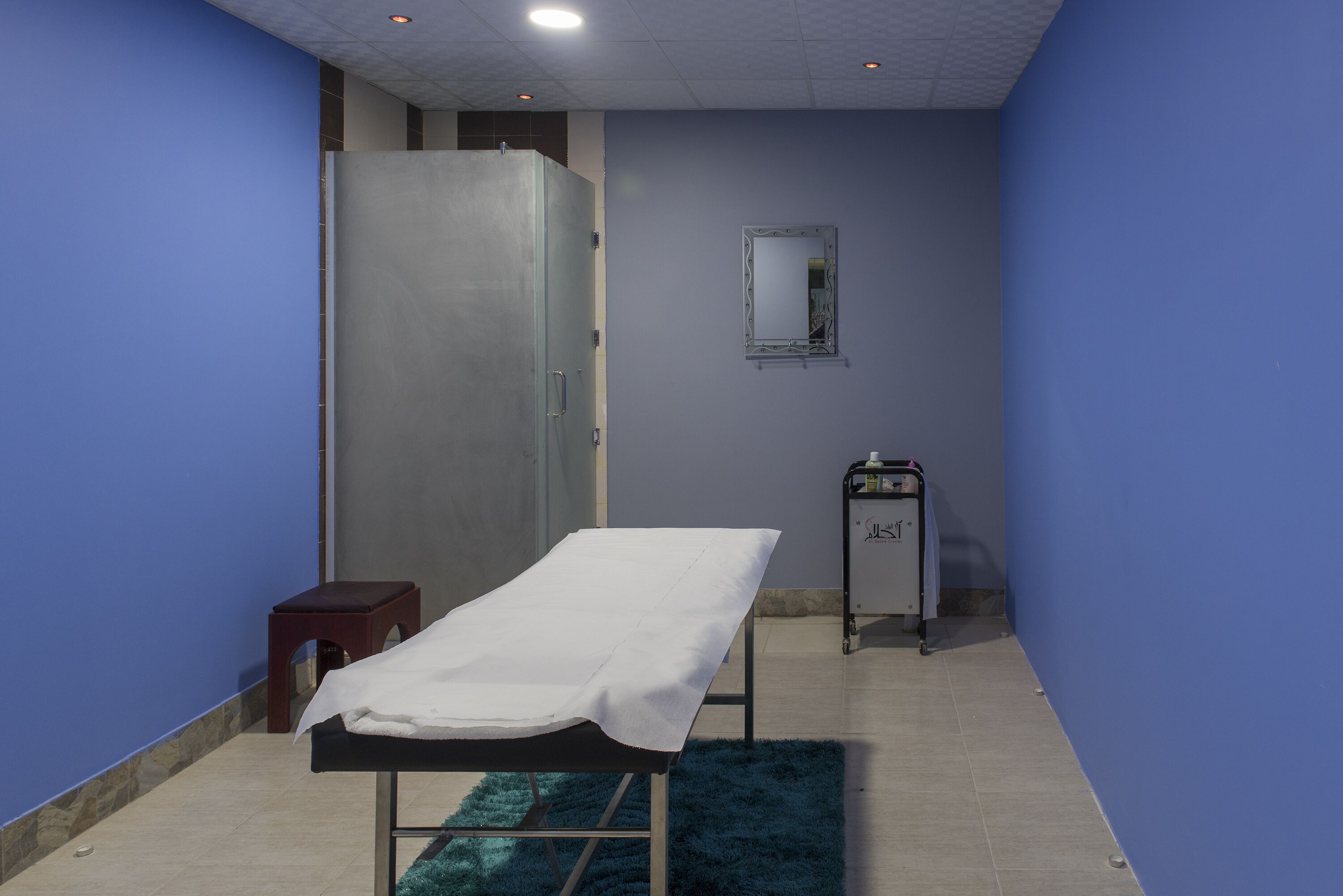 A clinical room with a single examination table, a small stool, a cabinet, and a mirror on the wall.