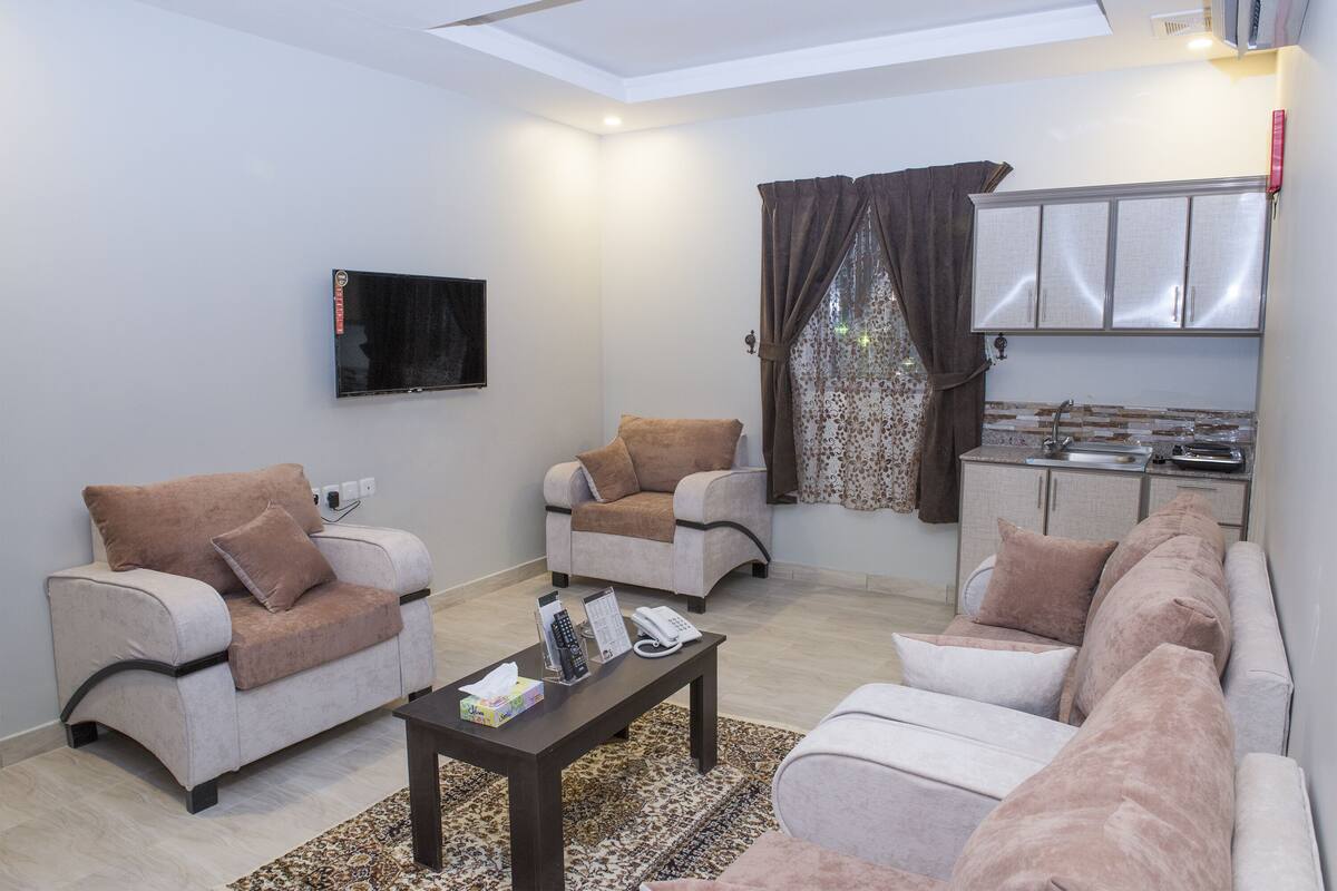Una sala de estar con dos sofás, una mesa de centro, un televisor y una zona de cocina con armarios y una ventana con cortinas.