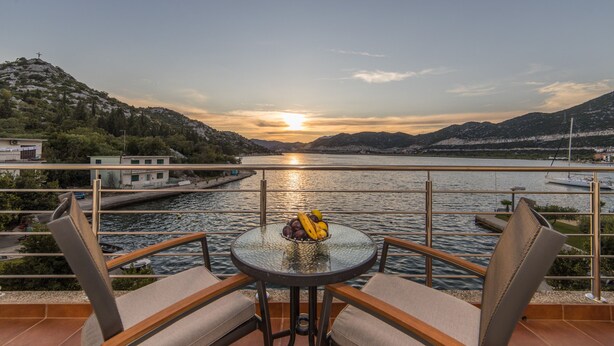 A balcony with a table and chairs, overlooking a body of water and hills during sunset. - Apartments Ema-Comfort Two Bedroom Apartment with Sea View (Rogotin)