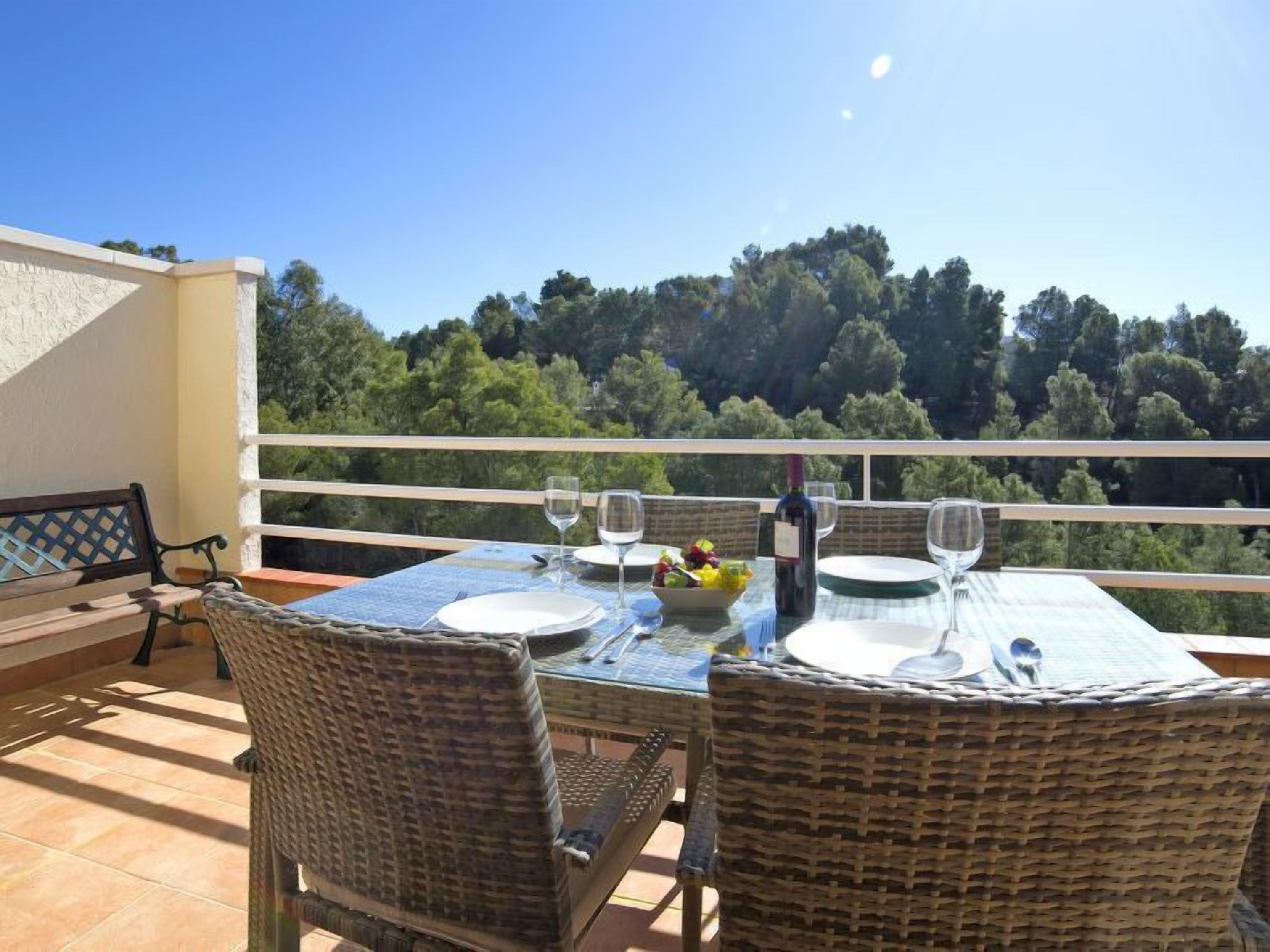 Terraço com cadeiras de vime e mesa posta para dois, com vista para uma área arborizada.