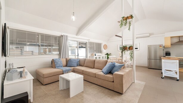 A modern living room with a beige sectional sofa, a white coffee table, and a dining area with a white table and chairs. There is a kitchen area with a stainless steel refrigerator and a white cabinet. - Little Kingsley Beach House - Back Flat - across the road from the Beach (Boat Harbour)