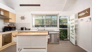 A modern kitchen with a central island, a built-in oven, a dishwasher, and a window with blinds. - Little Kingsley Beach House - Back Flat - across the road from the Beach (Boat Harbour)