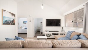 A modern living room with a beige sofa, a TV mounted on the wall, and a window with blinds. - Little Kingsley Beach House - Back Flat - across the road from the Beach (Boat Harbour)