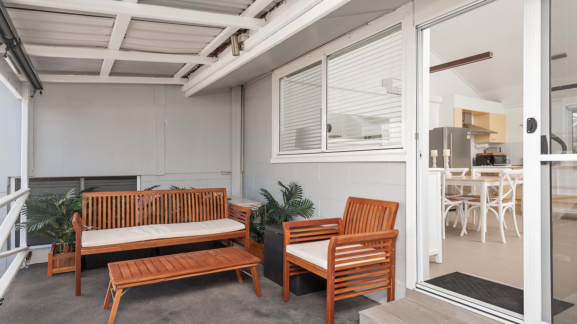 A balcony with wooden furniture, a sliding glass door leading to a kitchen, and a view of the building's interior.