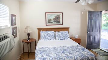 A bedroom with a bed, a bedside table with a lamp, a framed picture on the wall, and a door leading outside.