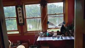 A room with a wooden floor, a desk with fishing equipment, a window overlooking a lake, and a sign reading "HANDMADE LURES INC." - Riverfront rental (South Elgin)