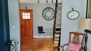 A room with a wooden floor, a wooden chair with a cushion, a small table, a clock, and a door leading to another room. - Riverfront rental (South Elgin)