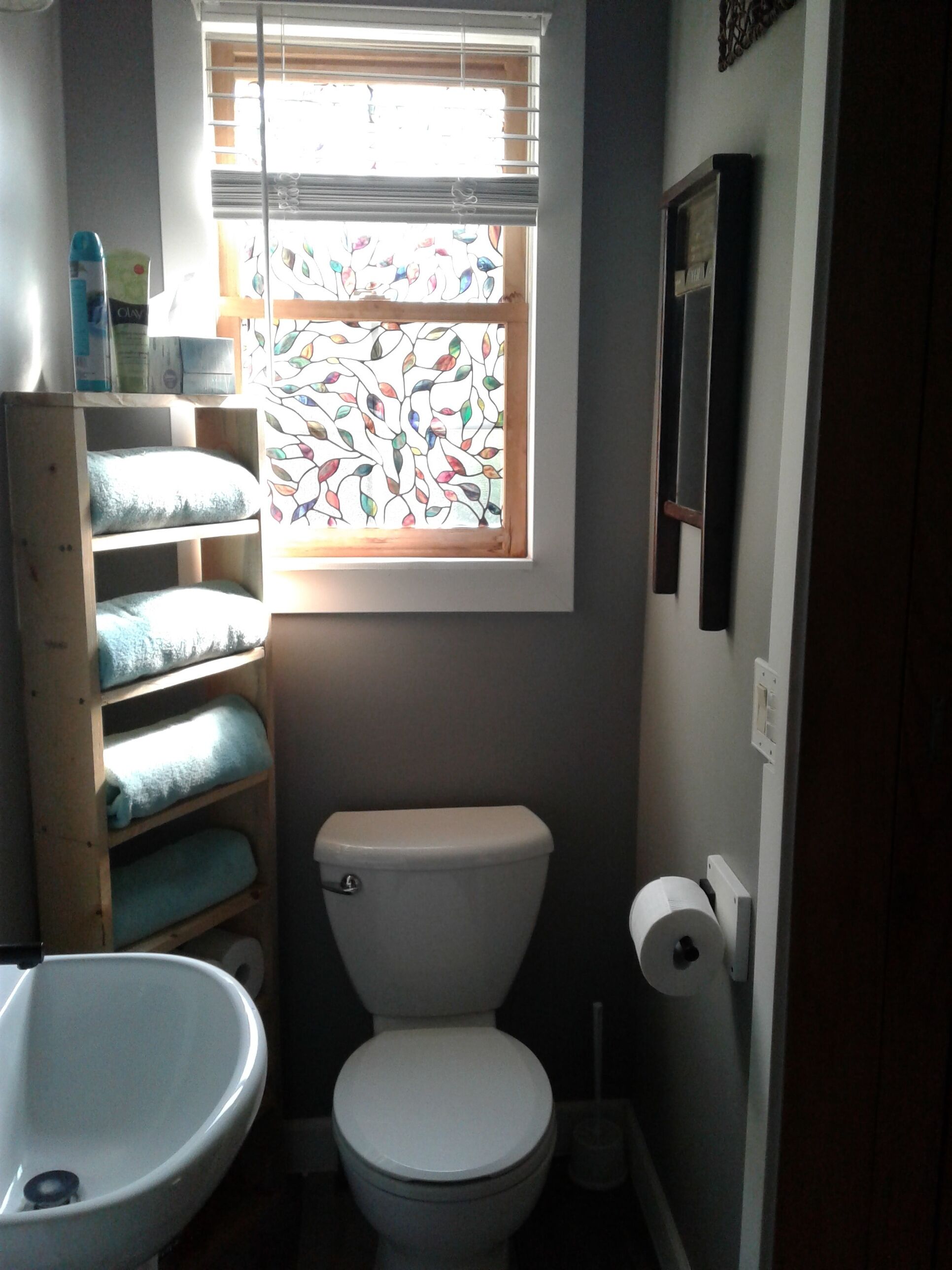 Une petite salle de bain avec des toilettes, une fenêtre ornée de vitraux et une étagère en bois où sont posées des serviettes.