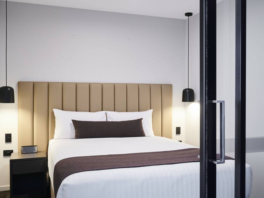 Un lit bien fait, avec une tête de lit, des tables de chevet et des lampes fixées au mur.