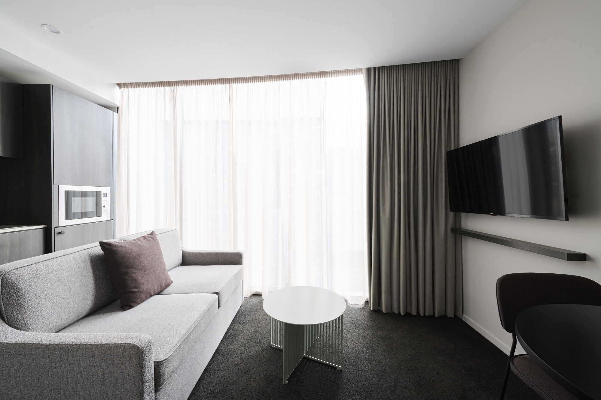 A modern hotel room with a grey sofa, a small round table, a flat-screen TV mounted on the wall, and a microwave built into the cabinetry.