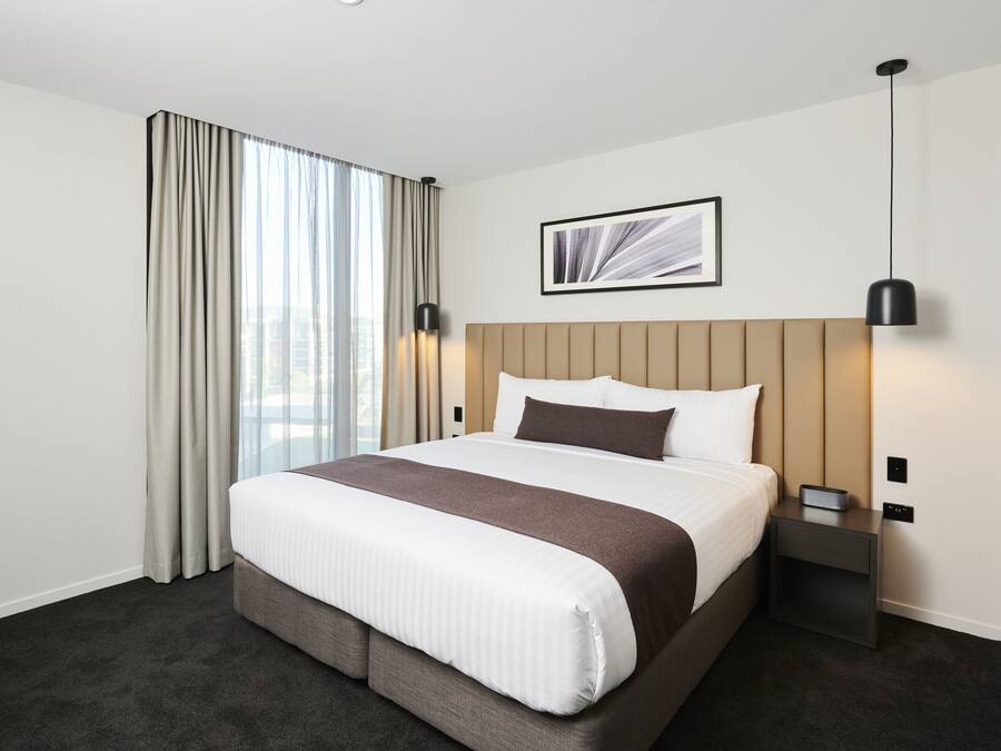A hotel room with a large bed, a headboard, a bedside table, a lamp, and a window with curtains.