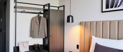 A hotel room with a bed, a wardrobe, a hanging coat rack, a bedside table with a cup and a book, and a pendant light.