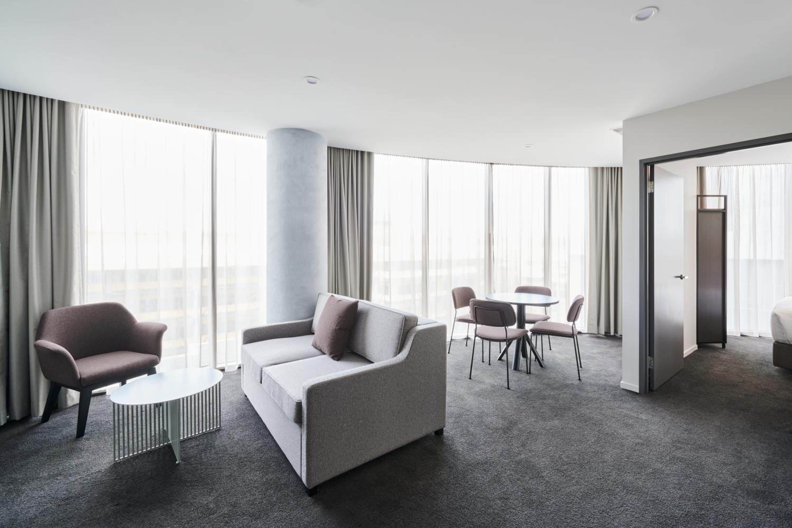 A modern hotel room with a grey sofa, a round coffee table, and a dining area with chairs and a table.
