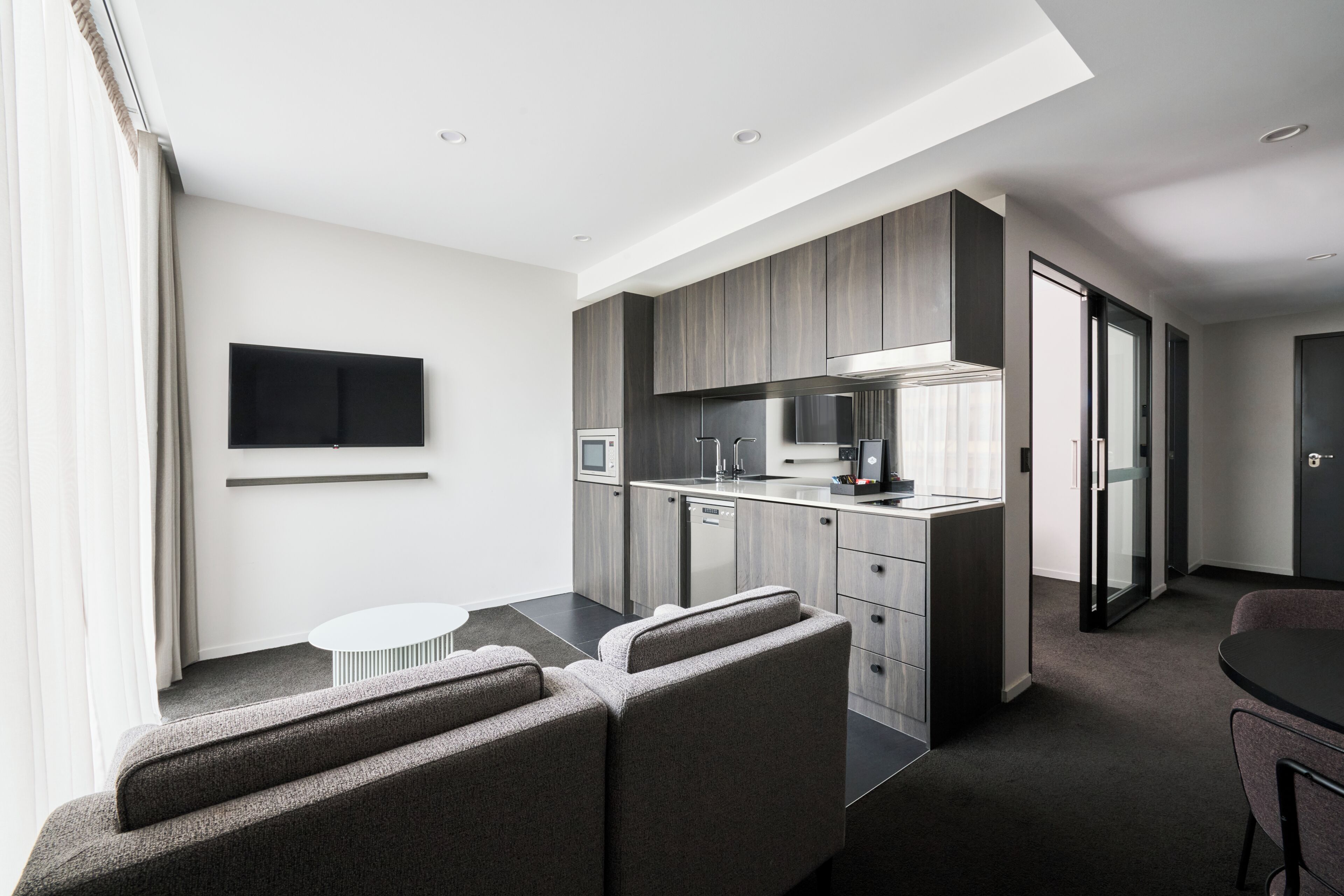A modern hotel room with a kitchenette, a flat-screen TV, a sofa, and a dining area.