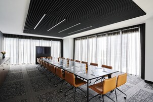 Un salón de conferencias con una mesa larga, sillas, un televisor de pantalla plana y ventanas amplias con cortinas.