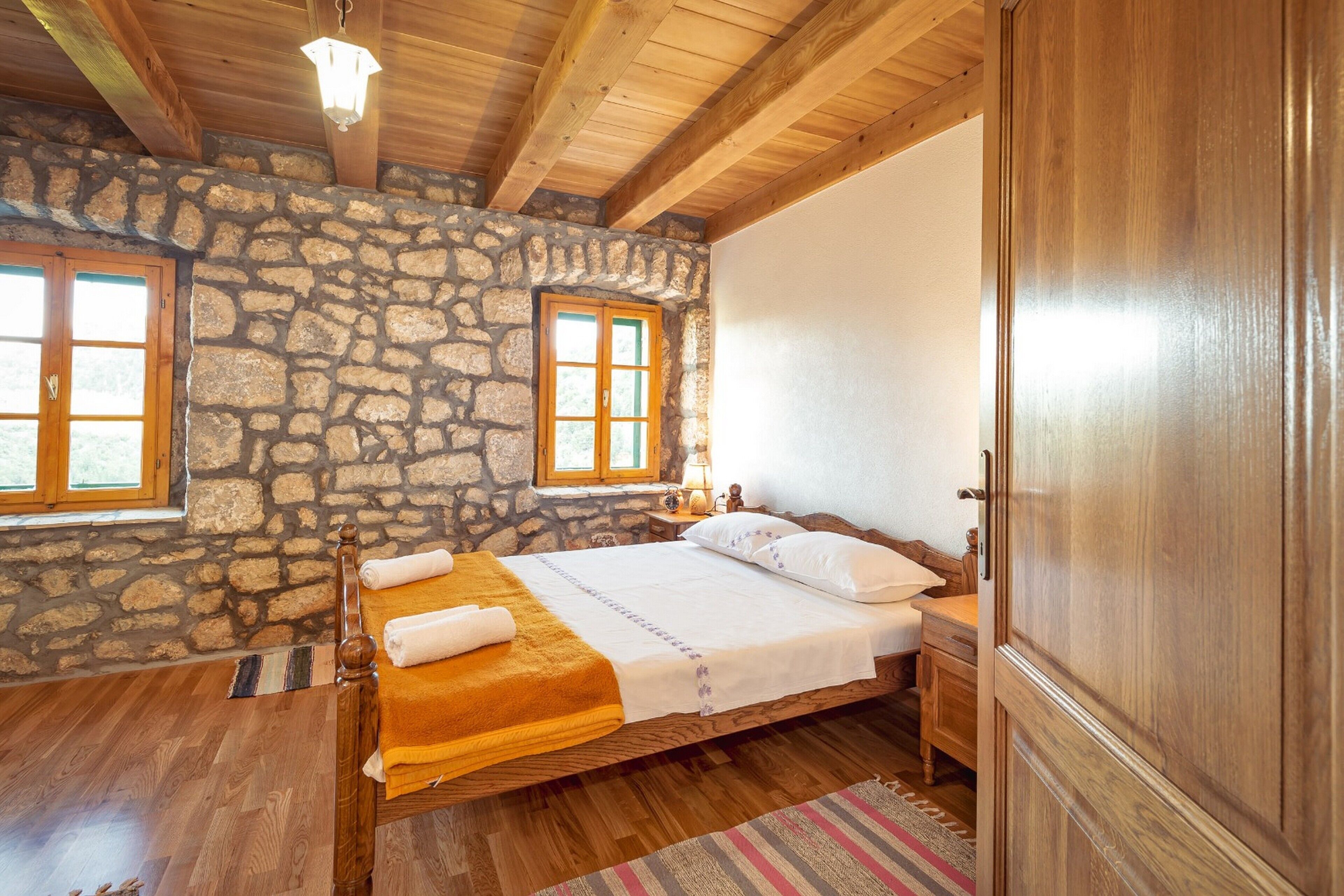 A bedroom with a stone wall, wooden ceiling beams, a bed with white linens, and a wooden wardrobe.