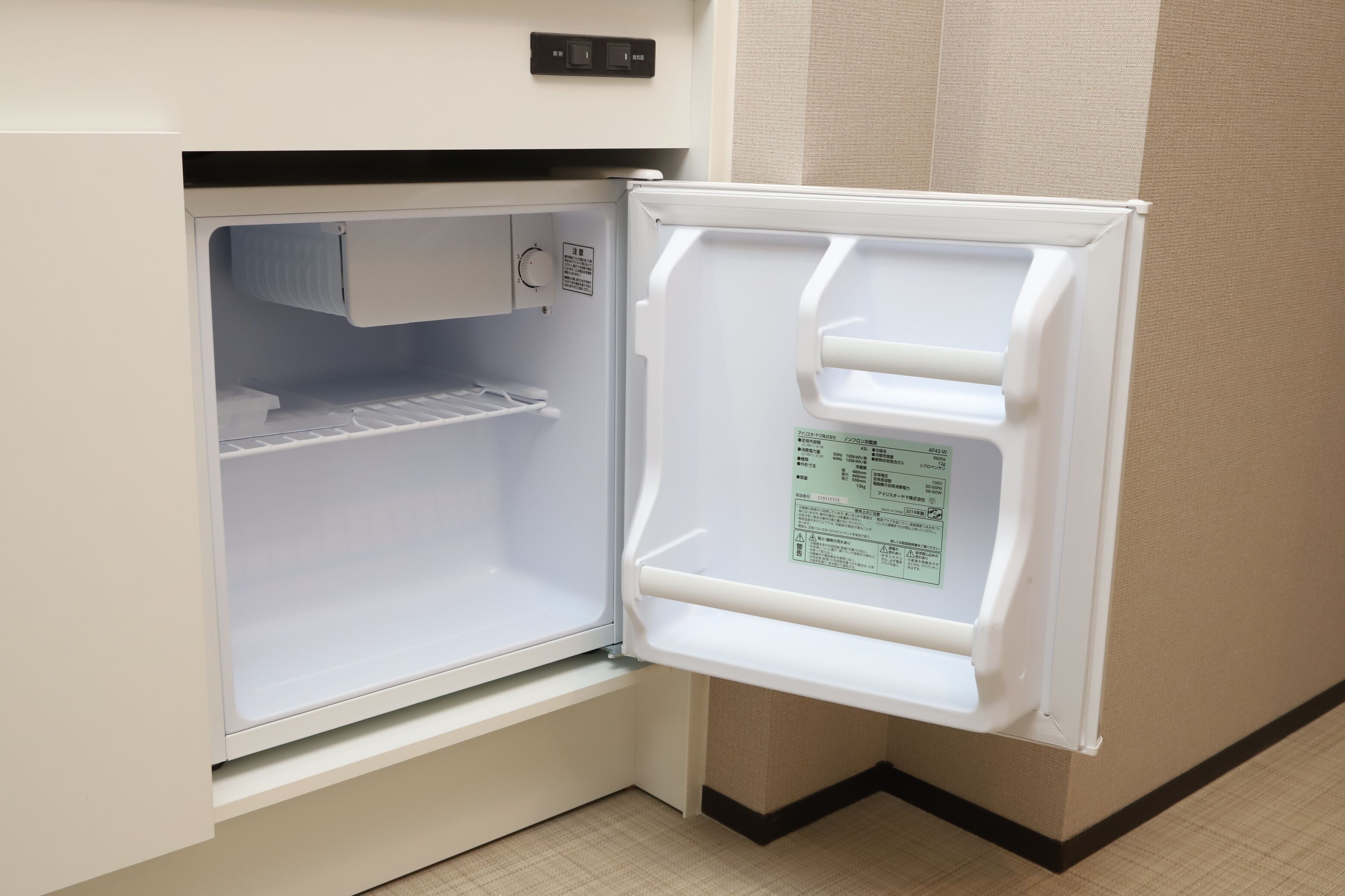 An open refrigerator with a control panel above it, located in a corner of a room with beige walls and a patterned floor.